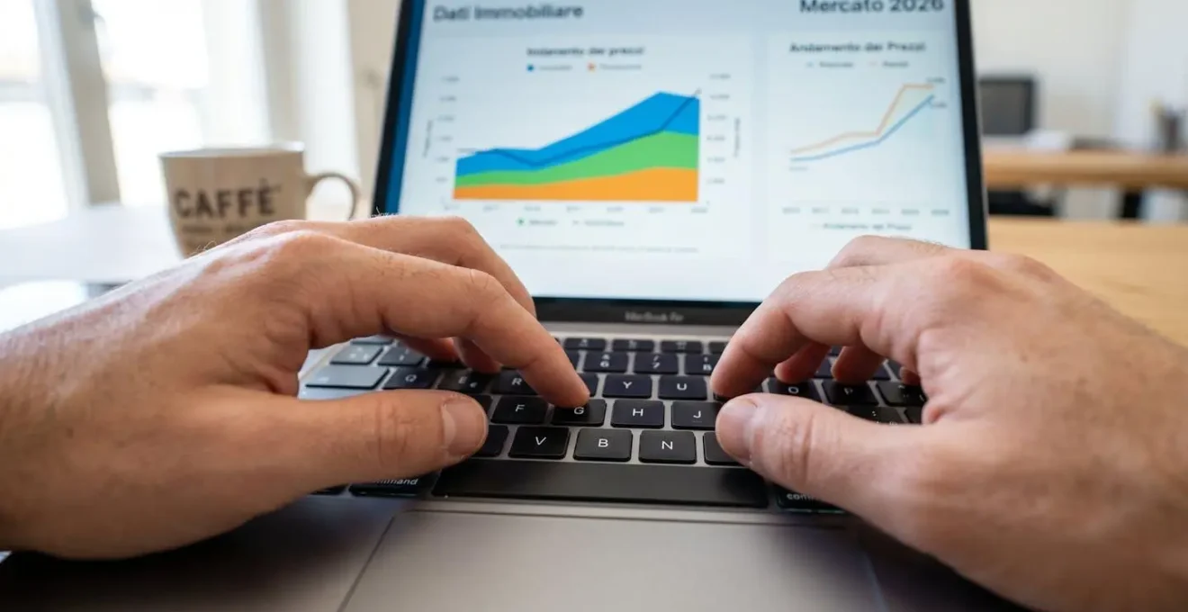 Primo piano ravvicinato di mani che digitano su una tastiera moderna di laptop con grafici immobiliari parzialmente visibili sullo schermo sfocato